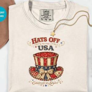 以下が含まれることがあります： クリーム色のTシャツ。赤、白、青の模様のシルクハットと「Hats Off to the USA」の文字がプリントされています。 金色のネックレスには星のペンダントが付いています。