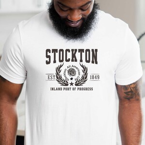 May include: White t-shirt with a Stockton graphic. The design includes the word "STOCKTON" above a circular emblem with text and the year 1849. The emblem is flanked by laurel branches and the text "INLAND PORT OF PROGRESS".