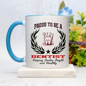 May include: White ceramic mug with a blue handle and rim, featuring the text "PROUD TO BE A DENTIST" above a cartoon tooth with dental tools. The design includes a black laurel wreath and the words "Keeping Smiles Bright and Healthy".