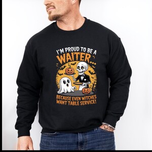 May include: A black long-sleeve sweatshirt featuring a graphic of a skeleton waiter holding a pumpkin on a tray next to a ghost. The text reads I'M PROUD TO BE A WAITER... BECAUSE EVEN WITCHES WANT TABLE SERVICE! in orange and white lettering.