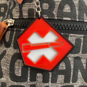 May include: A red and black keychain with a silver chain and key ring. The keychain has a geometric design with a white X and a bullet. The background is a black and gray patterned fabric.