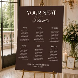 Puede incluir: Un cartel rectangular de color marrón oscuro para la distribución de asientos sobre un caballete de metal dorado. El cartel muestra texto blanco que dice "YOUR SEAT Awaits" seguido de nombres de invitados y "CELESTE'S BRIDAL SHOWER APRIL 18, 20XX."