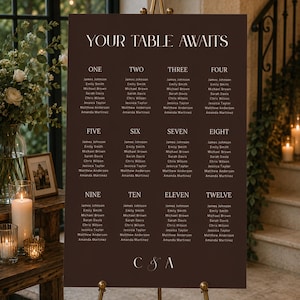 May include: A dark brown wedding seating chart sign with the text "YOUR TABLE AWAITS" at the top, followed by twelve numbered tables with lists of names underneath each, and the initials "C & A" printed in white at the bottom of the board.