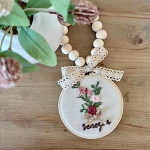 Könnte beinhalten: Ein kleiner, runder Stickrahmen mit beigefarbenem Stoffhintergrund. Der Rahmen zeigt ein handgesticktes Design eines Straußes aus rosa, roten und weißen Blumen mit grünen Blättern. Das Wort "forever" ist in braunem Garn unter den Blumen gestickt.