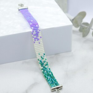 May include: A beaded bracelet with a gradient of purple, white and green beads. The bracelet has silver clasps.