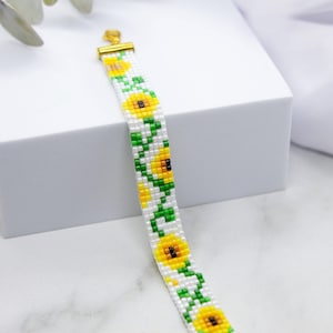 May include: A beaded bracelet featuring a sunflower design. The bracelet is white with yellow and green beads and has a gold clasp.