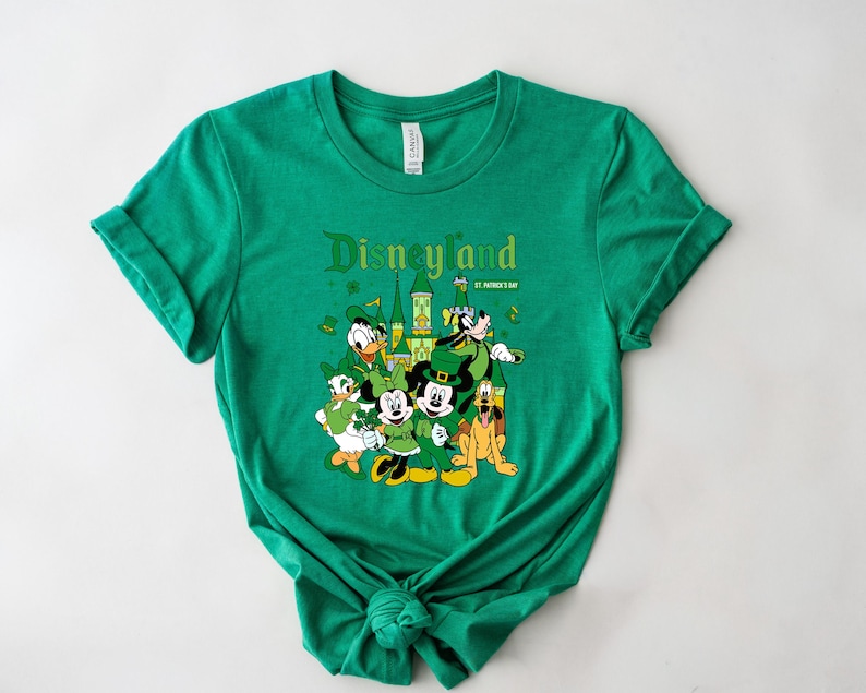 May include: Green t-shirt featuring a graphic of Disneyland characters dressed for St. Patrick's Day. The design includes Mickey Mouse, Donald Duck, Goofy, and Minnie Mouse, with the Disneyland logo.
