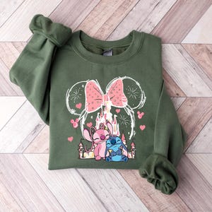 Peut inclure: Un sweat-shirt vert olive avec un motif de deux personnages de dessins anim&eacute;s, un rose et un bleu, avec un n&oelig;ud rose et un ch&acirc;teau. Le motif est soulign&eacute; en blanc avec des c&oelig;urs roses et des feux d'artifice.