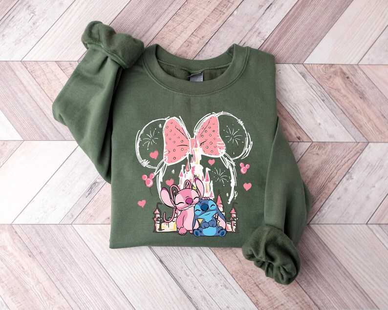 Peut inclure: Un sweat-shirt vert olive avec un motif de deux personnages de dessins anim&eacute;s, un rose et un bleu, avec un n&oelig;ud rose et un ch&acirc;teau. Le motif est soulign&eacute; en blanc avec des c&oelig;urs roses et des feux d'artifice.