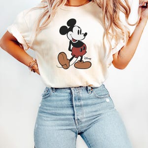 May include: A cream colored t-shirt with a black and white cartoon image of Mickey Mouse. The image shows Mickey Mouse walking with a smile and wearing red shorts with black buttons. The text 'Disney' is printed in black below the image.
