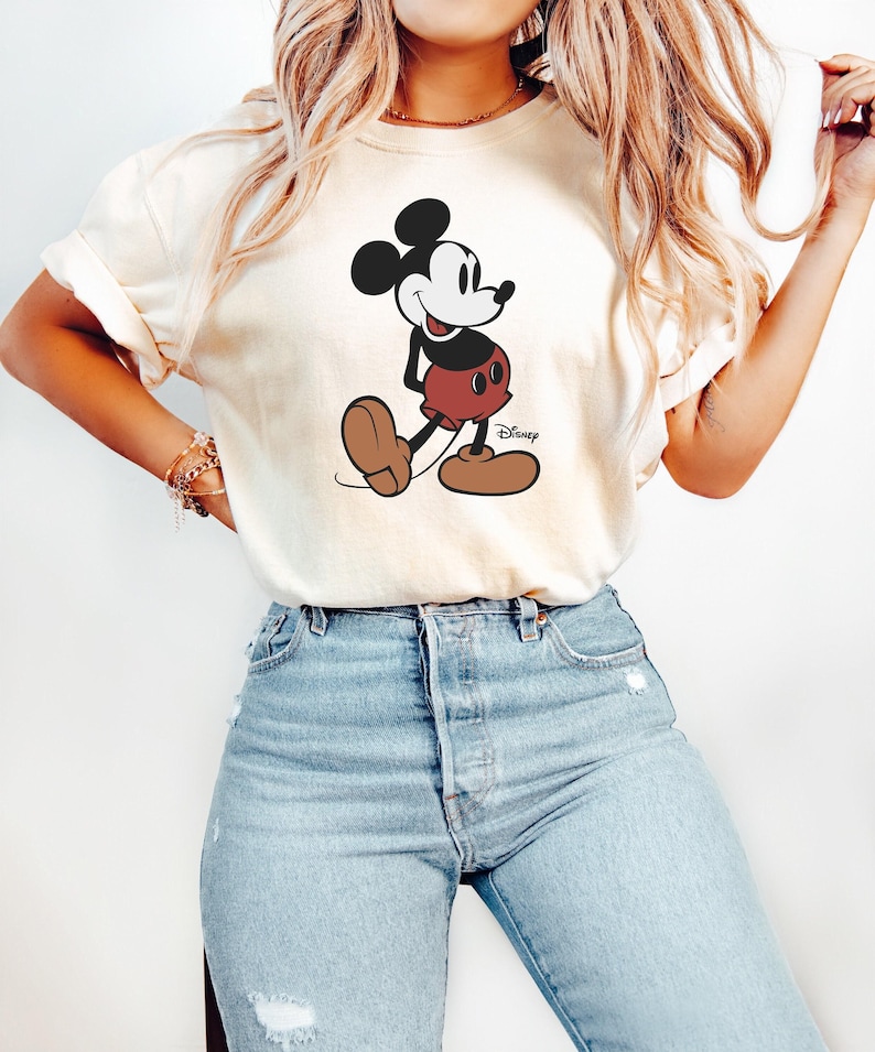May include: A cream colored t-shirt with a black and white cartoon image of Mickey Mouse. The image shows Mickey Mouse walking with a smile and wearing red shorts with black buttons. The text 'Disney' is printed in black below the image.
