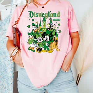 May include: Pink t-shirt featuring a Disneyland graphic with Mickey Mouse, Donald Duck, and other Disney characters in St. Patrick's Day attire. The text "Disneyland" is in green, with "St. Patrick's Day" below.