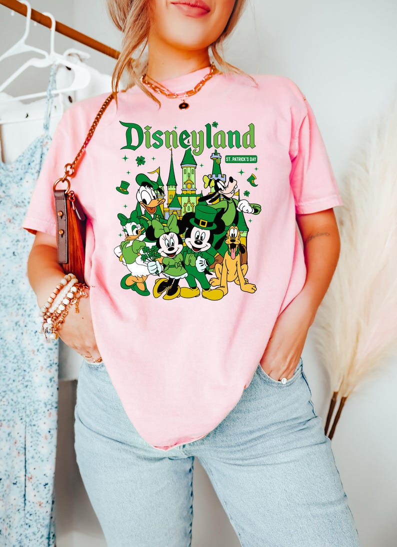 May include: Pink t-shirt featuring a Disneyland graphic with Mickey Mouse, Donald Duck, and other Disney characters in St. Patrick's Day attire. The text "Disneyland" is in green, with "St. Patrick's Day" below.