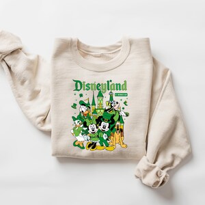May include: A cream-colored sweatshirt featuring a graphic of Disneyland characters dressed for St. Patrick's Day. The design includes Mickey and Minnie Mouse, Donald Duck, Goofy, and Pluto, with the Disneyland logo in green.