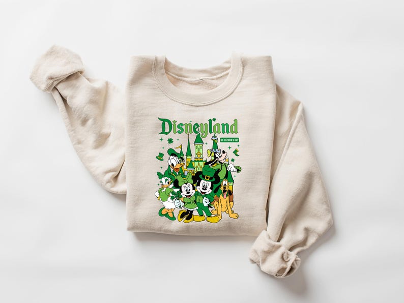 May include: A cream-colored sweatshirt featuring a graphic of Disneyland characters dressed for St. Patrick's Day. The design includes Mickey and Minnie Mouse, Donald Duck, Goofy, and Pluto, with the Disneyland logo in green.