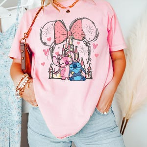 Peut inclure: T-shirt rose clair avec un motif de ch&acirc;teau, d'un n&oelig;ud et de deux personnages de dessin anim&eacute;. Le motif comprend des c&oelig;urs et des feux d'artifice. Le t-shirt est de couleur rose clair.