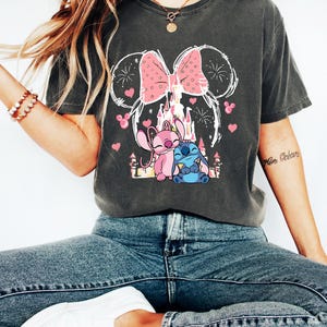 Peut inclure: T-shirt gris fonc&eacute; avec un motif d'un n&oelig;ud rose, des oreilles de souris et un ch&acirc;teau. En dessous, deux personnages de dessins anim&eacute;s, un rose et un bleu, avec des c&oelig;urs roses. Le t-shirt est de style d&eacute;contract&eacute;.
