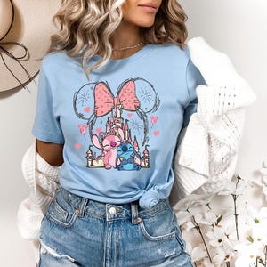 Peut inclure: T-shirt bleu clair avec un motif de ch&acirc;teau, d'un n&oelig;ud rose et de personnages de dessins anim&eacute;s. Le motif comprend un personnage rose et bleu, avec des c&oelig;urs et des feux d'artifice. Le t-shirt est &agrave; col rond et &agrave; manches courtes.