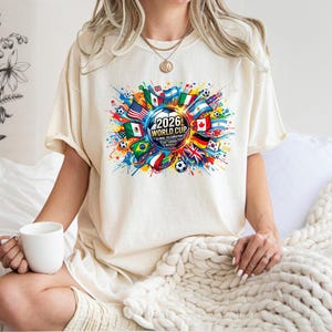May include: Ivory-colored t-shirt featuring a colorful graphic for the 2026 World Cup. The design includes flags, soccer balls, and the text "2026 World Cup Global Celebration". The shirt is a casual, short-sleeved style.