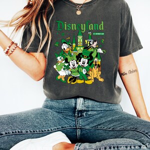May include: Dark gray t-shirt featuring a Disneyland graphic with Disney characters dressed for St. Patrick's Day. The design includes Mickey and Minnie Mouse, Donald Duck, Goofy, and a castle. The shirt is worn with blue jeans and white sneakers.