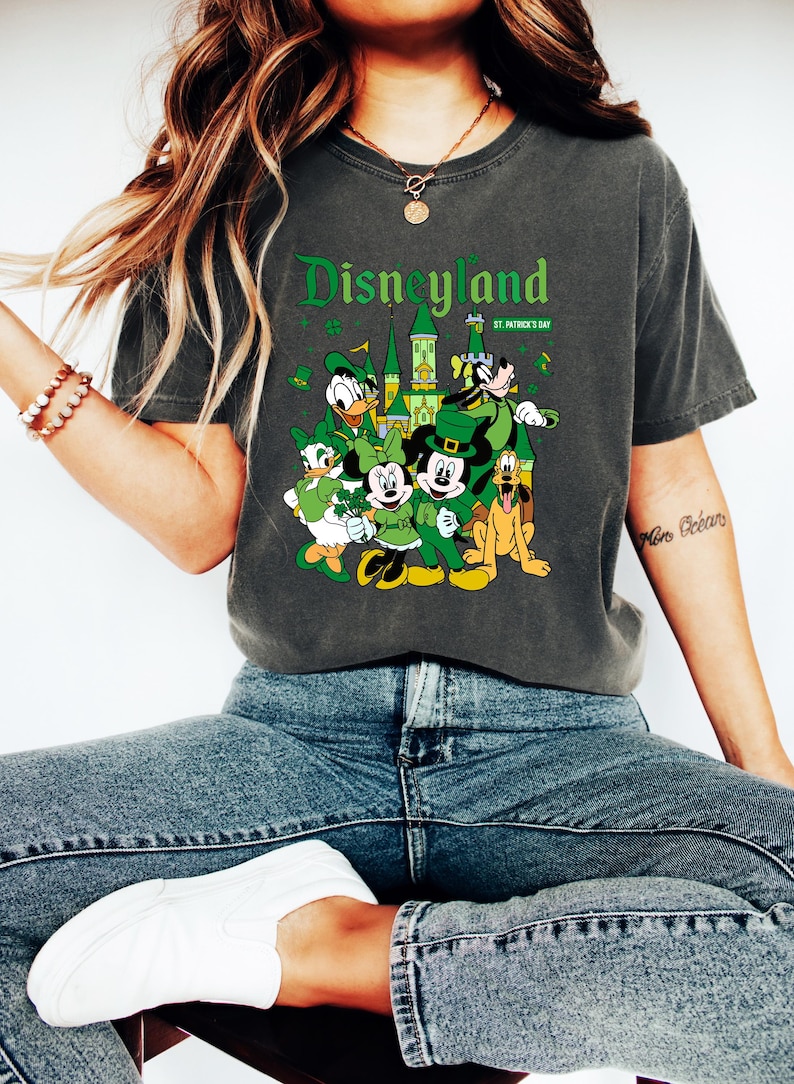 May include: Dark gray t-shirt featuring a Disneyland graphic with Disney characters dressed for St. Patrick's Day. The design includes Mickey and Minnie Mouse, Donald Duck, Goofy, and a castle. The shirt is worn with blue jeans and white sneakers.