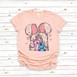 Peut inclure: T-shirt rose clair avec un motif de ch&acirc;teau, deux personnages de dessins anim&eacute;s et un grand n&oelig;ud. Les personnages sont roses et bleus. Le t-shirt a un col rond et des manches courtes.