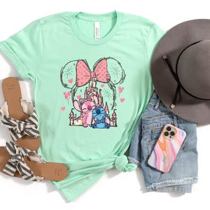 Peut inclure: Un t-shirt vert menthe avec un motif des personnages Disney Stitch et Angel, un n&oelig;ud rose et un ch&acirc;teau. Le t-shirt est pr&eacute;sent&eacute; avec des sandales, un short en jean, un chapeau gris et une coque de t&eacute;l&eacute;phone.