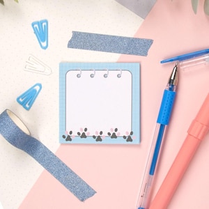 May include: A blue notepad with a white page and a row of paw prints along the bottom. The notepad has a spiral binding and is sitting on a pink surface. There are two blue pens and a blue paper clip on the surface.