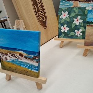 Puede incluir: Tres pequeñas pinturas sobre caballetes. Las pinturas son de una escena de playa, un patrón floral y un paisaje. Los caballetes son de madera y están pintados en un color madera natural.