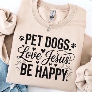 May include: A beige sweatshirt with the text "PET DOGS Love Jesus BE HAPPY" in black. The design includes paw prints, hearts, and sunbursts. A silver necklace is visible near the neckline. The sweatshirt is paired with blue jeans.