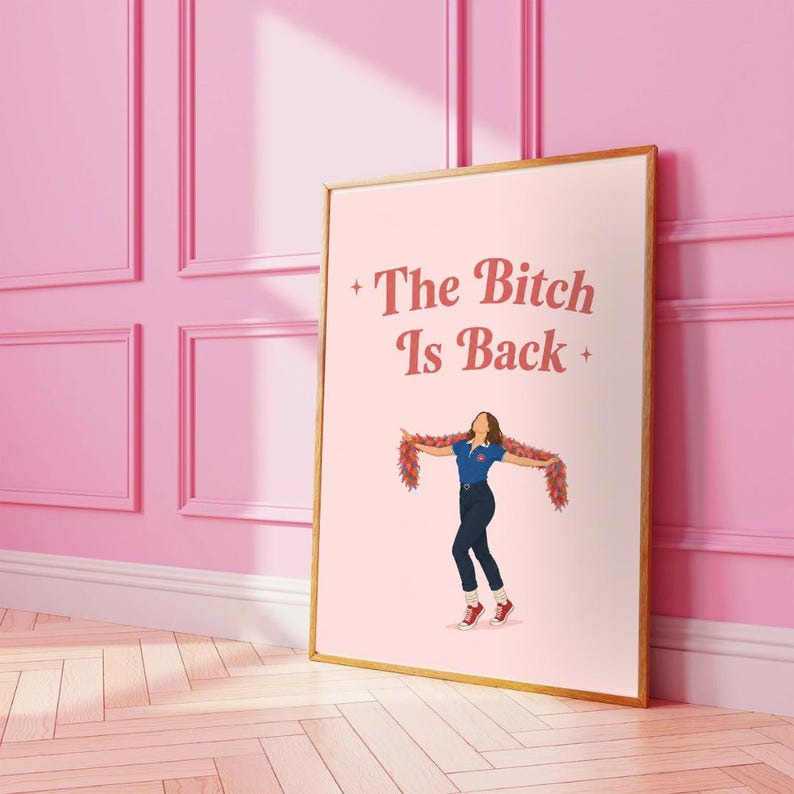 Może przedstawiać: Oprawiony wydruk artystyczny z r&oacute;żowym tłem i złotą ramą. Wydruk zawiera tekst "The Bitch Is Back" w stylizowanej czcionce. Ilustracja osoby w niebieskiej koszuli, ciemnych spodniach i czerwonych butach znajduje się na środku.