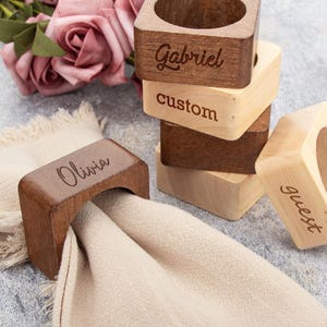 Könnte beinhalten: Serviettenringe aus Holz mit eingravierten Namen und dem Wort "Gast". Die Ringe sind in verschiedenen Braun- und Naturholztönen gehalten, mit einer beigen Serviette in einem. Ein Stapel Ringe ist sichtbar.