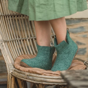 Può includere: Un paio di stivali in feltro verde acqua con un design a punta unico. Gli stivali sono indossati da una persona seduta su una sedia di vimini con un cuscino marrone. La persona indossa una gonna verde.