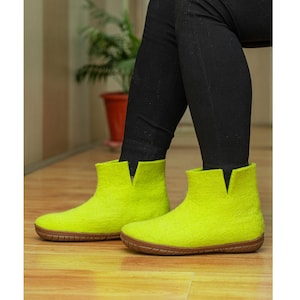 Può includere: Un paio di stivaletti in lana infeltrita color verde lime brillante, caratterizzati da un design minimalista senza lacci, con una distinta tacca a V al centro del colletto e una suola in gomma marrone flessibile e testurizzata.