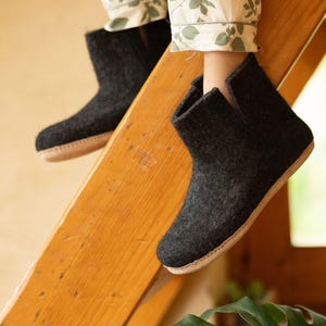 May include: Dark grey felted wool ankle boots with a light brown sole. The boots are shown on a wooden staircase. These boots are a cosy and comfortable footwear option for indoor use. The boots are a stylish and practical choice for anyone looking for warm and comfortable footwear.