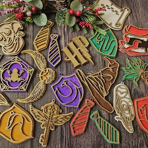 Könnte beinhalten: Eine Sammlung von Holzornamenten zum Thema Harry Potter, darunter Krawatten, Schilde und das Hogwarts-Logo. Die Ornamente weisen goldene, violette, grüne, rote und braune Akzente mit einer goldenen Glitzerkontur auf. Die Dekorationen sind auf einer Holzoberfläche angeordnet.