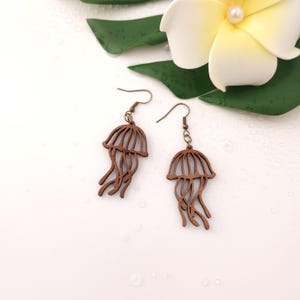 Puede incluir: Un par de pendientes de madera con forma de medusa. Los pendientes son de color marr&oacute;n c&aacute;lido, con un dise&ntilde;o detallado que imita la forma de una medusa. Los pendientes cuelgan de alambres para las orejas de color bronce. Una flor blanca y una hoja verde est&aacute;n en el fondo.