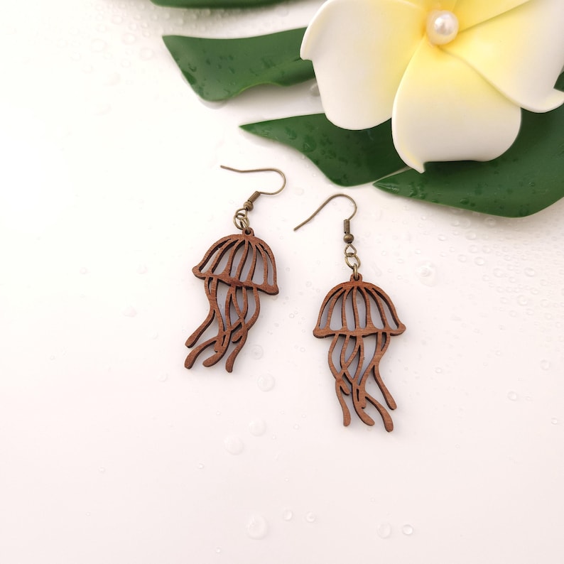 Puede incluir: Un par de pendientes de madera con forma de medusa. Los pendientes son de color marr&oacute;n c&aacute;lido, con un dise&ntilde;o detallado que imita la forma de una medusa. Los pendientes cuelgan de alambres para las orejas de color bronce. Una flor blanca y una hoja verde est&aacute;n en el fondo.