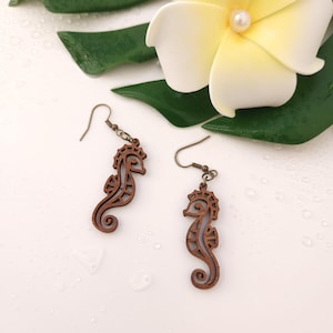Puede incluir: Un par de pendientes de madera con forma de caballito de mar con ganchos de color bronce. Los pendientes son de color marr&oacute;n oscuro y tienen un dise&ntilde;o calado detallado. Una flor blanca y hojas verdes est&aacute;n en el fondo.