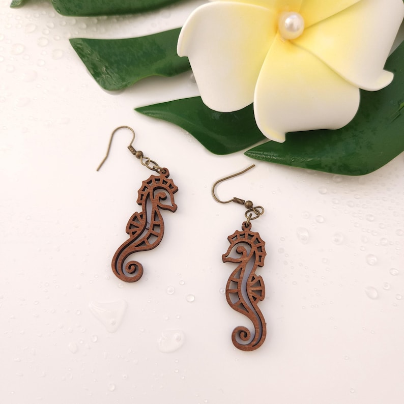 Puede incluir: Un par de pendientes de madera con forma de caballito de mar con ganchos de color bronce. Los pendientes son de color marr&oacute;n oscuro y tienen un dise&ntilde;o calado detallado. Una flor blanca y hojas verdes est&aacute;n en el fondo.