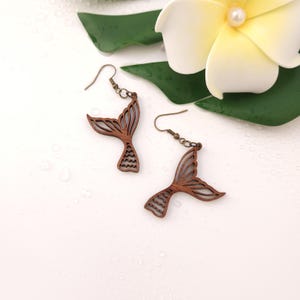 Puede incluir: Pendientes de cola de sirena de madera con ganchos de color bronce. Los pendientes son de color marr&oacute;n claro y tienen un dise&ntilde;o calado detallado. Sobre un fondo blanco, con una flor y hojas verdes.