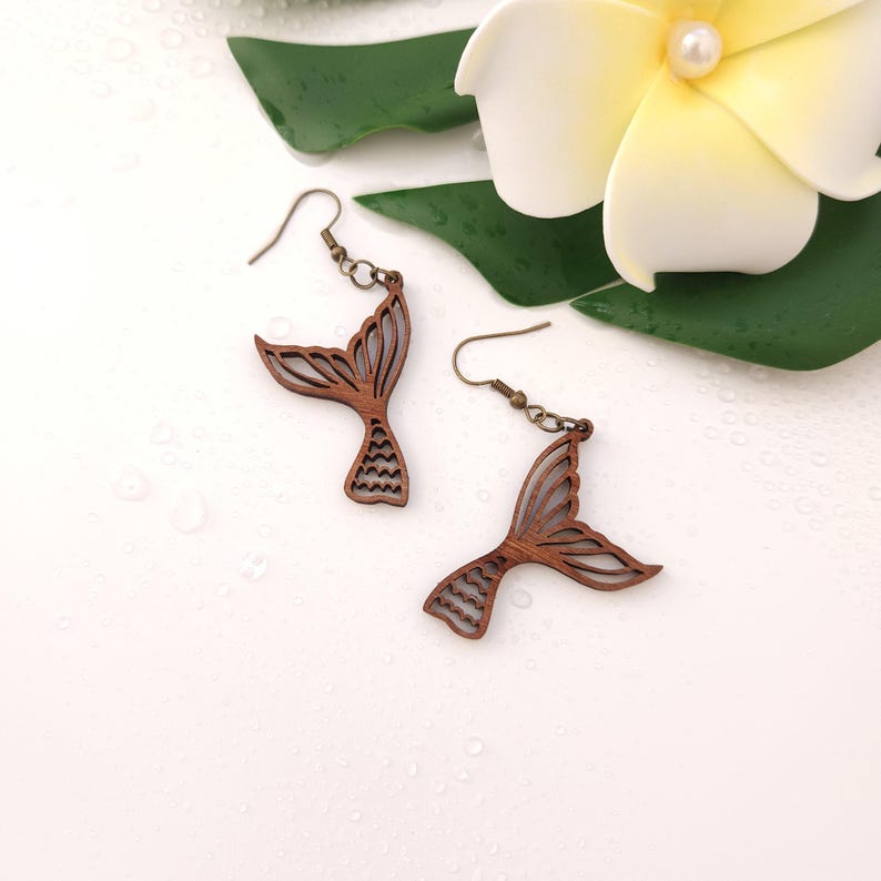 Puede incluir: Pendientes de cola de sirena de madera con ganchos de color bronce. Los pendientes son de color marr&oacute;n claro y tienen un dise&ntilde;o calado detallado. Sobre un fondo blanco, con una flor y hojas verdes.