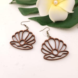 Puede incluir: Un par de pendientes de madera con dise&ntilde;o de concha. Los pendientes son de color marr&oacute;n c&aacute;lido y tienen un dise&ntilde;o recortado. Los pendientes cuelgan de alambres para las orejas de color bronce. Sobre una superficie blanca con una flor y hojas verdes.
