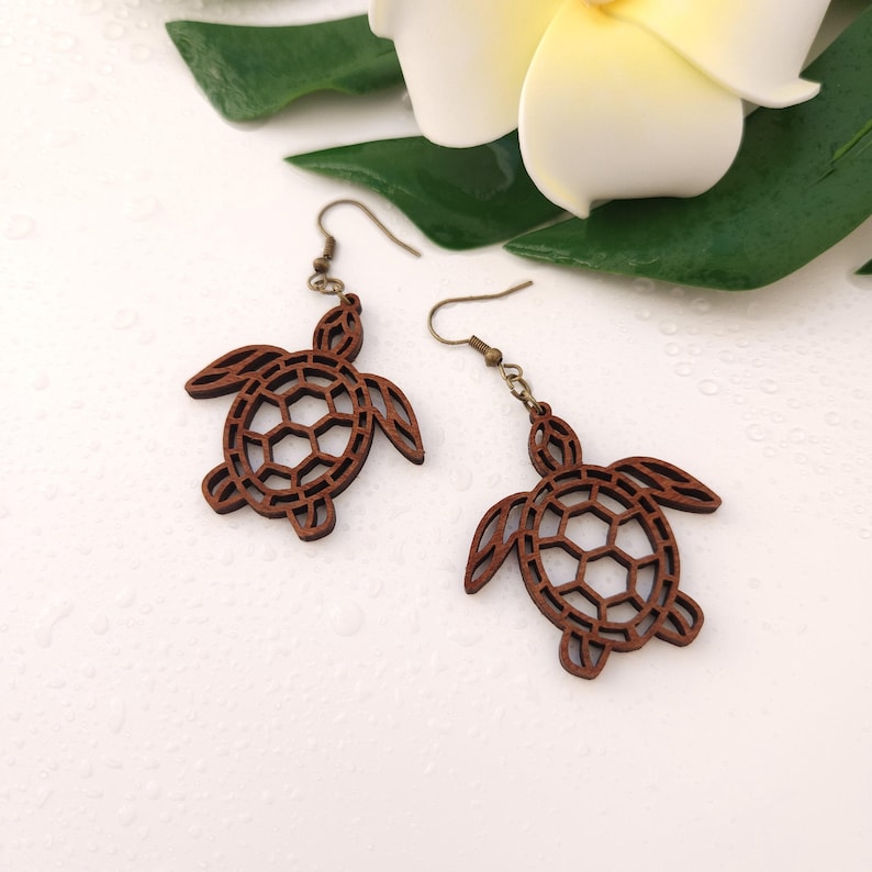 Puede incluir: Un par de pendientes de madera con forma de tortuga. Los pendientes son marrones con un dise&ntilde;o recortado, que muestra el caparaz&oacute;n y el cuerpo de la tortuga. Tienen cierre de gancho y se exhiben sobre una superficie blanca.