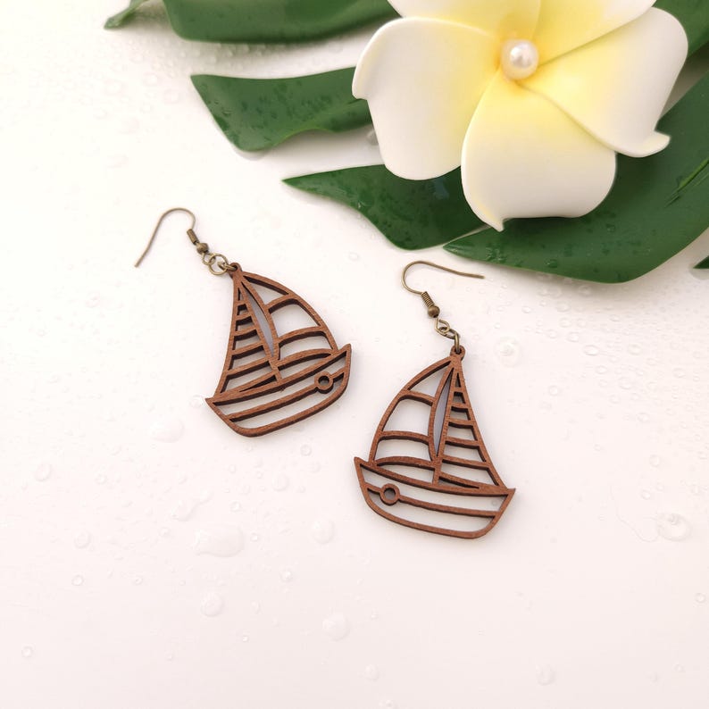 Puede incluir: Un par de pendientes de madera con forma de velero y ganchos de color bronce. Los pendientes son de color marr&oacute;n claro y presentan un dise&ntilde;o recortado de un velero. Se muestran sobre una superficie blanca con una flor y hojas verdes.