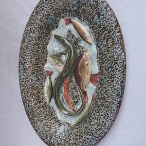Op de afbeelding: Ovale keramische wandkunst met een reliëf van verschillende vissen, waaronder een paling, in bruin, groen en rood. De achtergrond heeft een gespikkeld, getextureerd patroon in bruin en blauw. Een decoratief stuk voor een nautisch of kustthema.