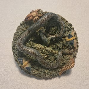 Könnte beinhalten: Eine detaillierte, kreisförmige Skulptur mit einer gewundenen Schlange mit einem strukturierten, grau-silbernen Muster. Die Skulptur umfasst eine Krabbe, Schmetterlinge und andere Insekten, allesamt auf einem Bett aus grünem Laub. Das Gesamtdesign hat ein naturalistisches Thema.