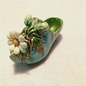 Op de afbeelding: Een keramisch decoratief object in de vorm van een schoen, geschilderd in turquoise en groen. Een grote witte madelief met een gele kern en groene bladeren siert de voorkant. Een decoratief item in vintage-stijl.