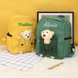 May include: Two personalized backpacks, one yellow and one green, each with a teddy bear in a pocket. The yellow backpack has the name "Matthew" embroidered, and the green backpack has "Alexis". Both have black accents.