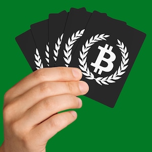 May include: A hand holding a fan of black playing cards with a white Bitcoin symbol encircled by a laurel wreath. The cards are against a solid green background. The Bitcoin symbol is a stylized letter "B".